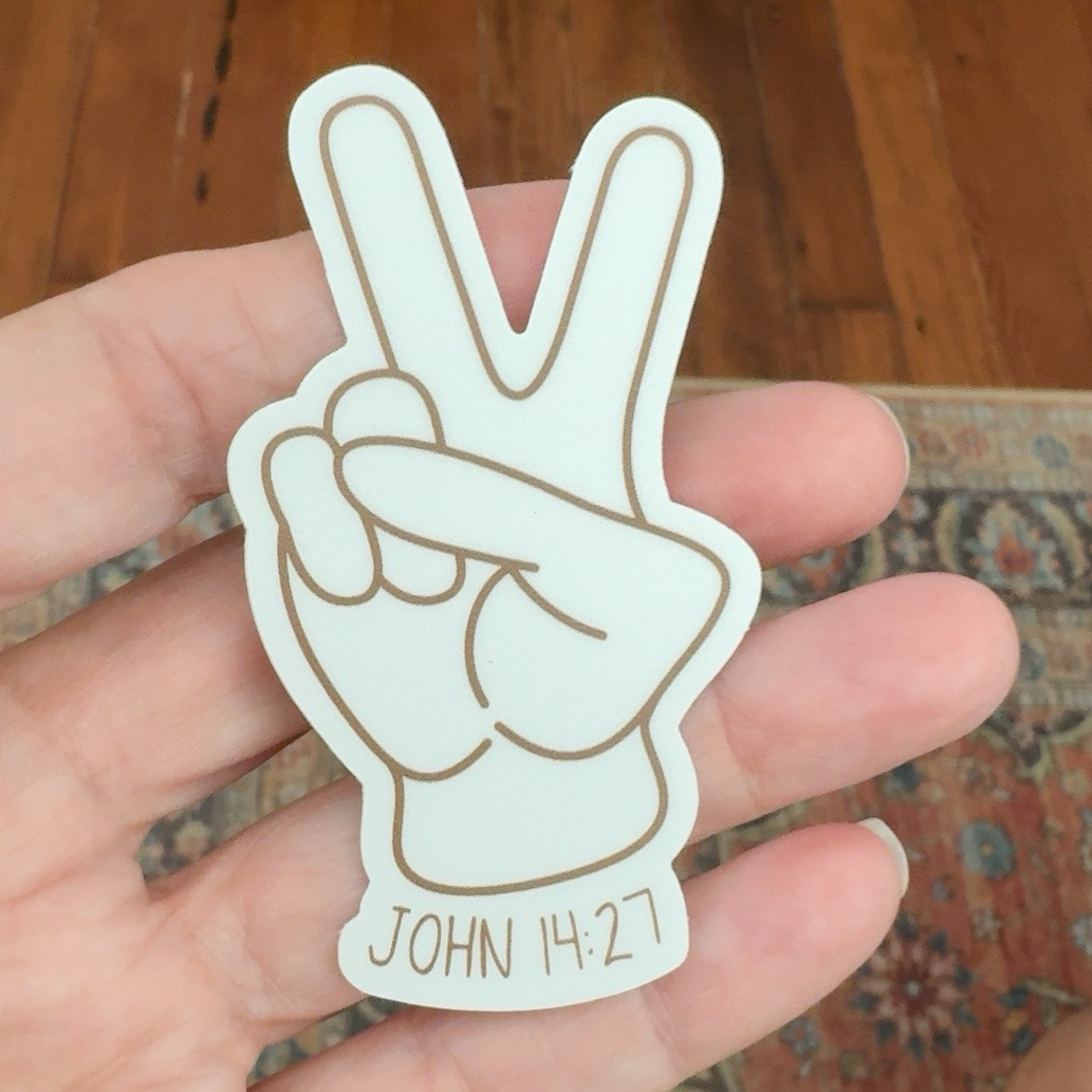 A hand holding a white die-cut sticker shaped like a peace hand gesture with brown outline details and the text 'JOHN 14:27' at the bottom, set against a background of a wooden floor and patterned rug.
