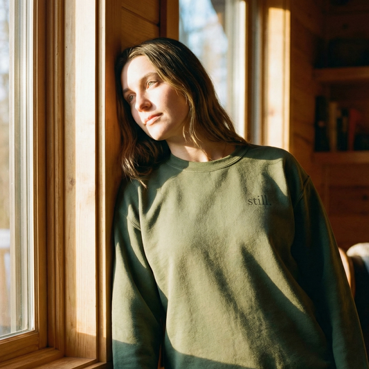 Minimalist green crewneck sweatshirt with the word “still.” worn in warm window light.