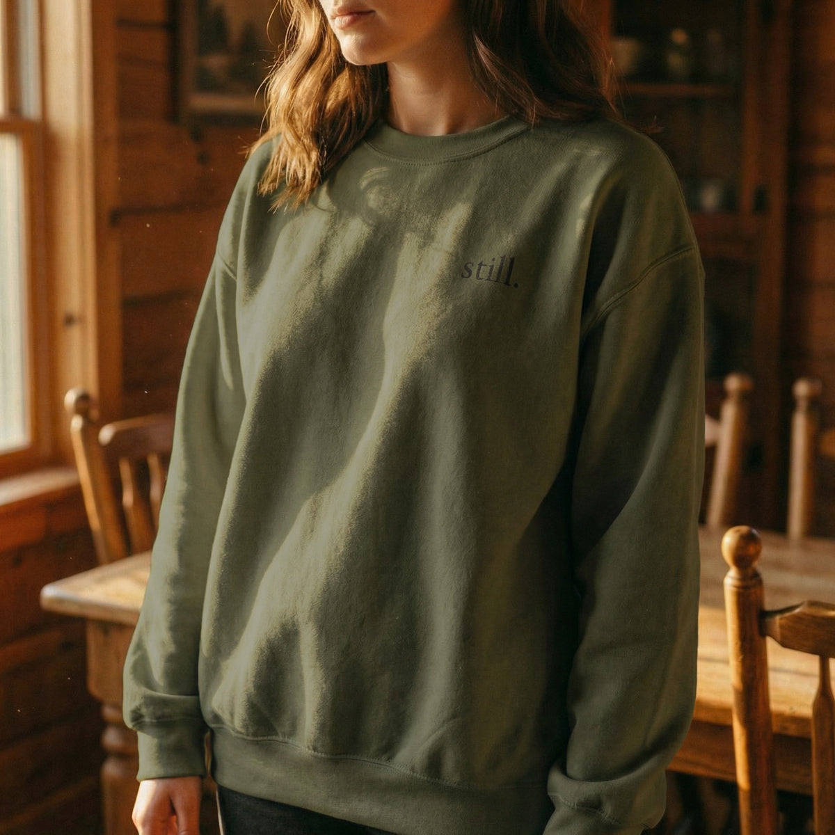 Green minimalist crewneck sweatshirt featuring the word “still.” on the chest in warm natural light.