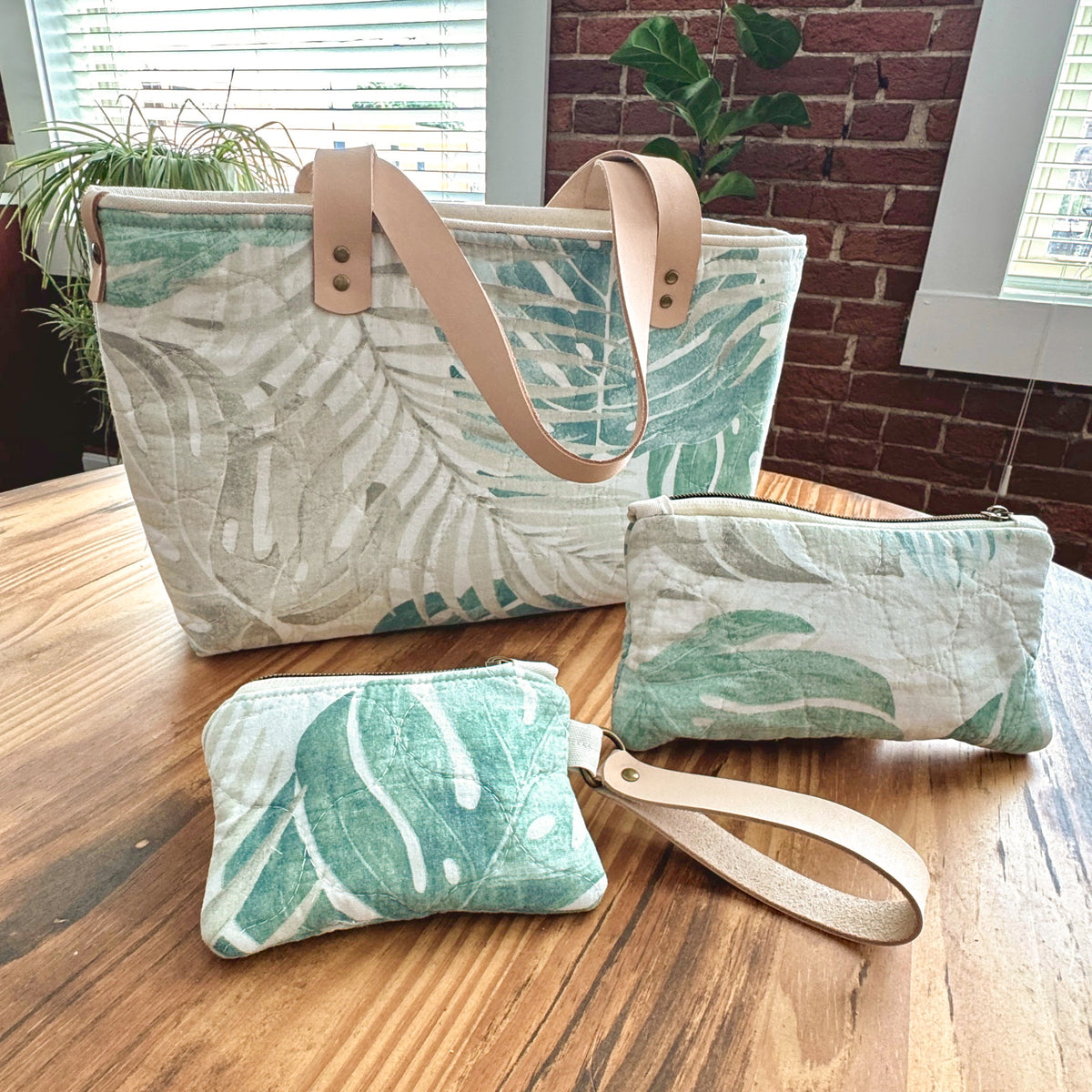A matching fabric tote bag and two small pouches with a tropical leaf print in green and beige, displayed on a wooden table in a home setting.