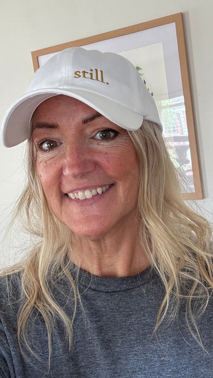 Woman wearing a white embroidered “still.” baseball hat in soft natural light, casual everyday outfit, calm and minimal style