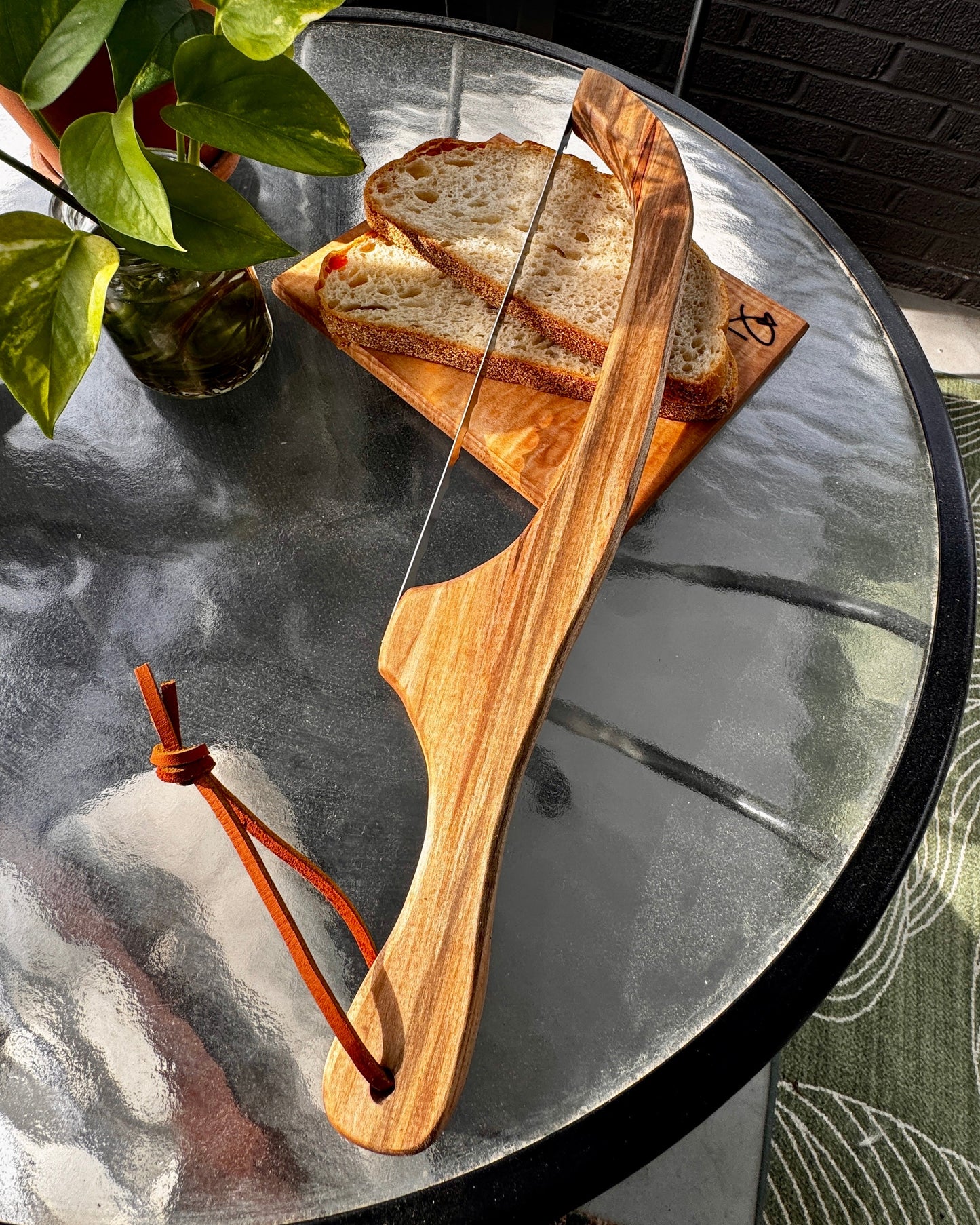 Handcrafted hardwood cutting board and knife with bread on a glass table next to a plant
