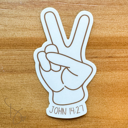 A white die-cut sticker featuring a hand making the peace sign with the text 'JOHN 14:27' at the bottom, placed on a wooden surface.