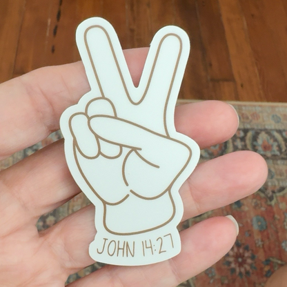 A hand holding a white die-cut sticker shaped like a peace hand gesture with brown outline details and the text 'JOHN 14:27' at the bottom, set against a background of a wooden floor and patterned rug.