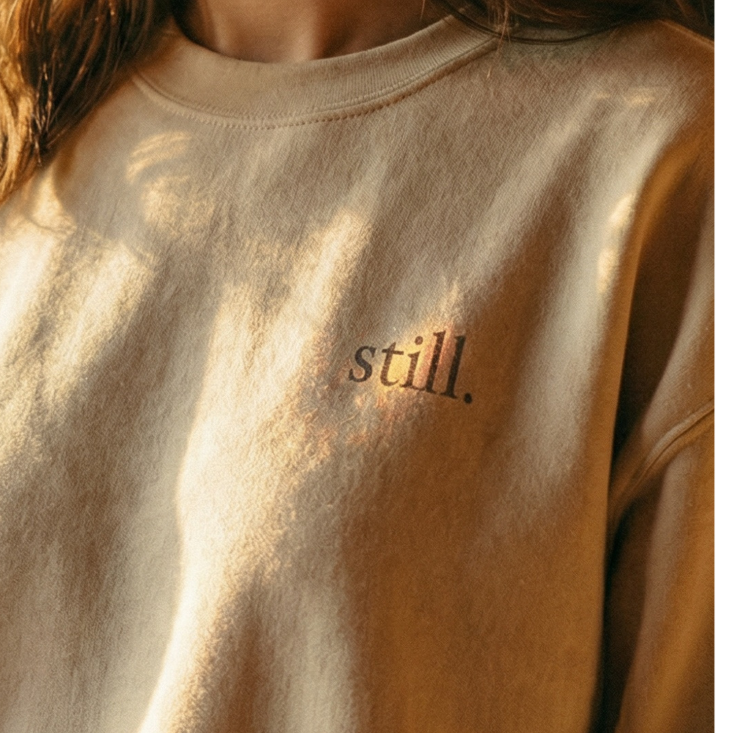 Minimalist neutral crewneck sweatshirt with the word “still.” printed on the chest in warm natural light.