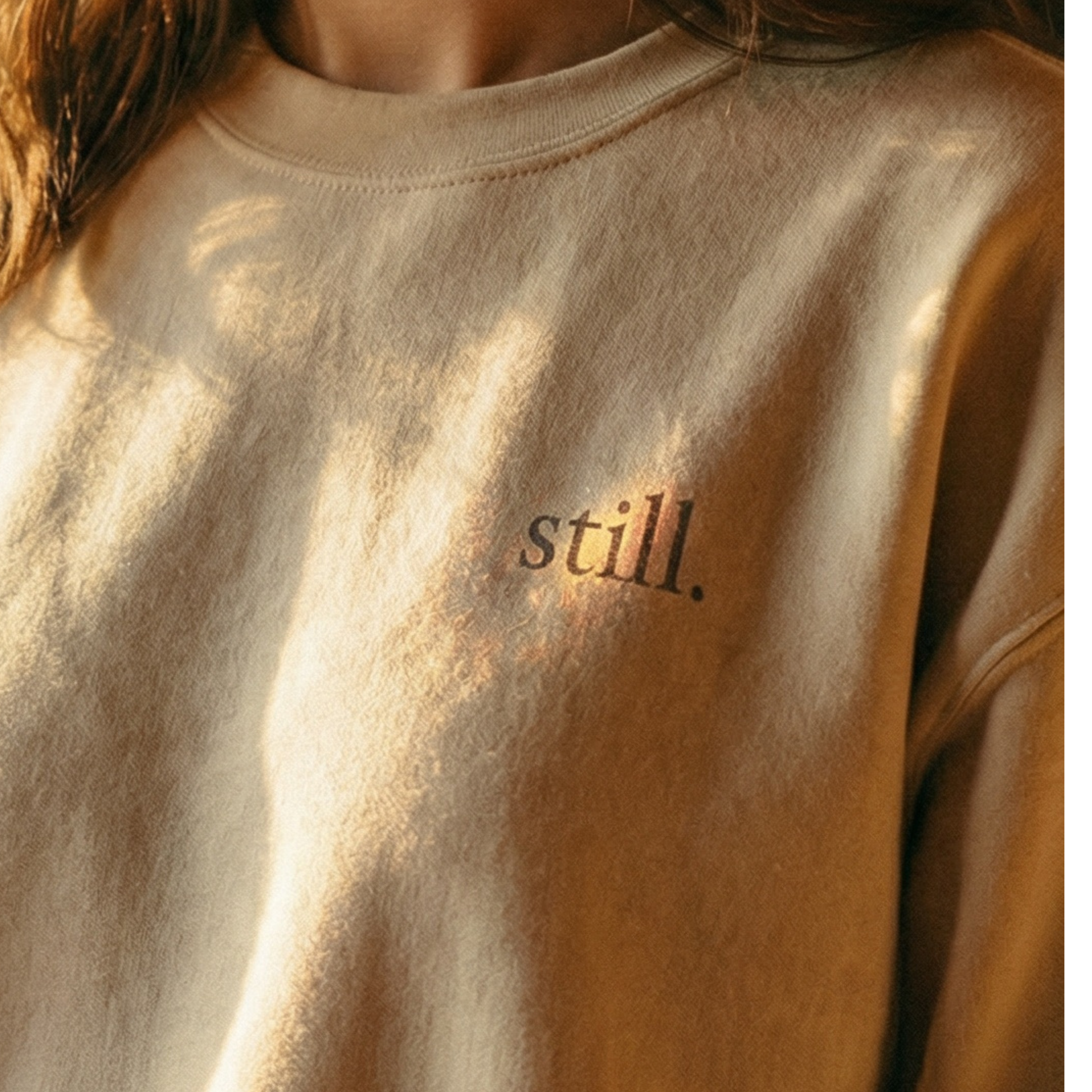 Minimalist neutral crewneck sweatshirt with the word “still.” printed on the chest in warm natural light.