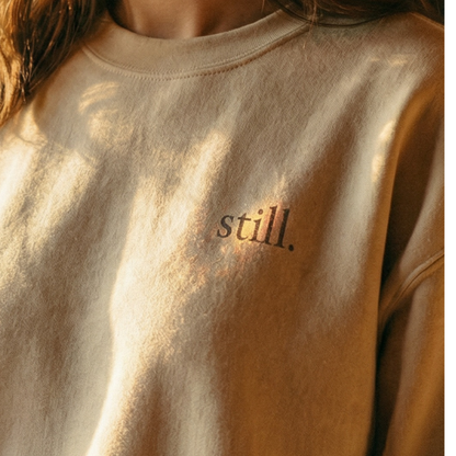 Minimalist neutral crewneck sweatshirt with the word “still.” printed on the chest in warm natural light.