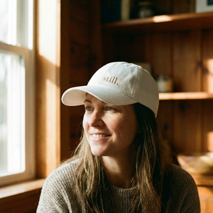 Woman wearing a cap with 'still.' text, sitting in a warm-lit room.