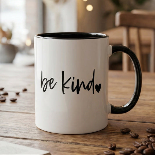 White mug with 'be kind' text on a wooden table with coffee beans and an open book.
