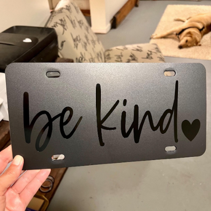 Black Be Kind plastic license plate with subtle black lettering and heart detail held in studio