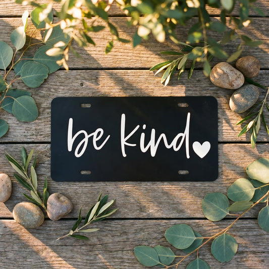 Black Be Kind plastic license plate with white script lettering and heart detail on wood background