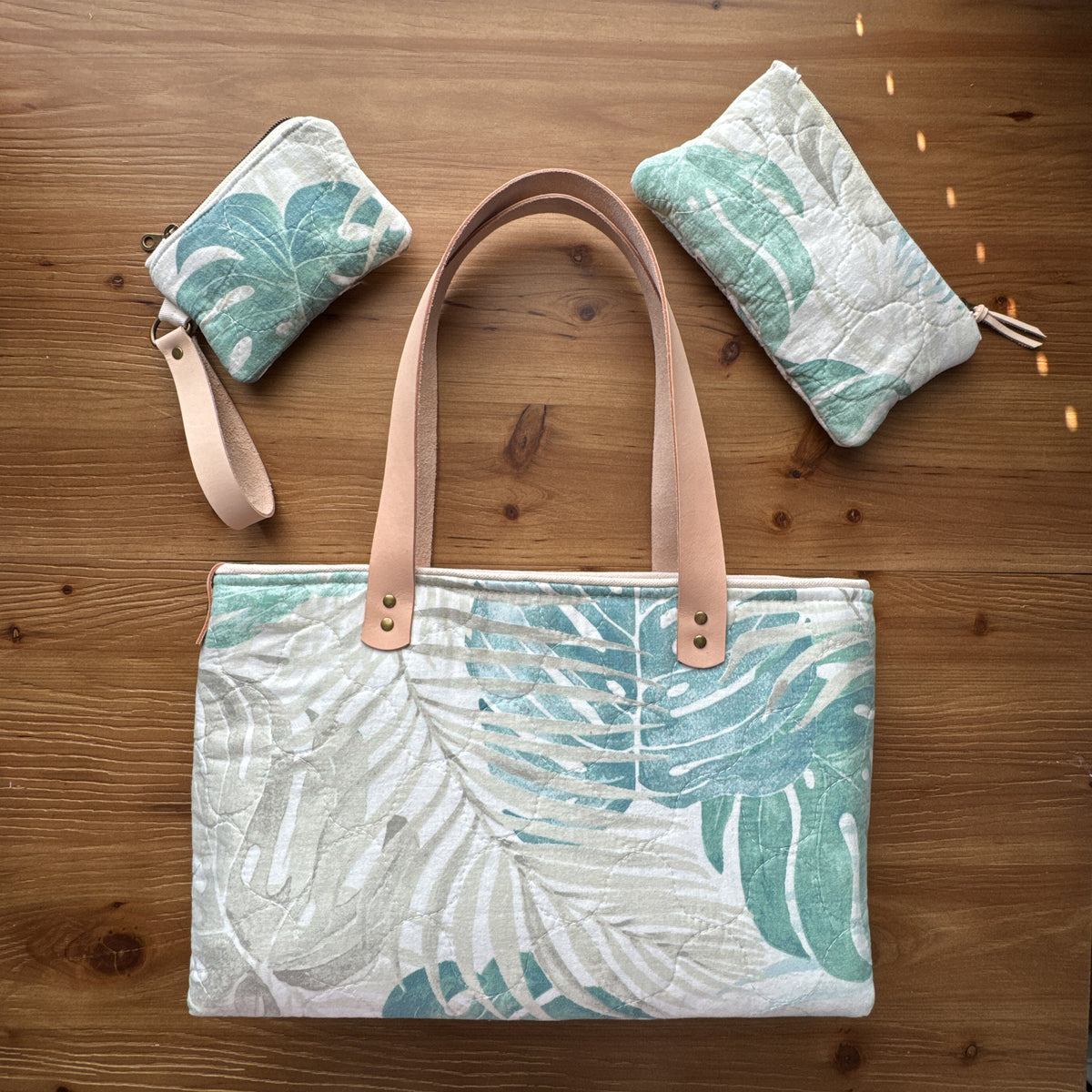 A tote bag with two matching zip pouches arranged on a wooden surface. The bag and pouches feature a white fabric printed with large teal and mint green tropical leaf patterns, with light tan leather handles and wrist strap.