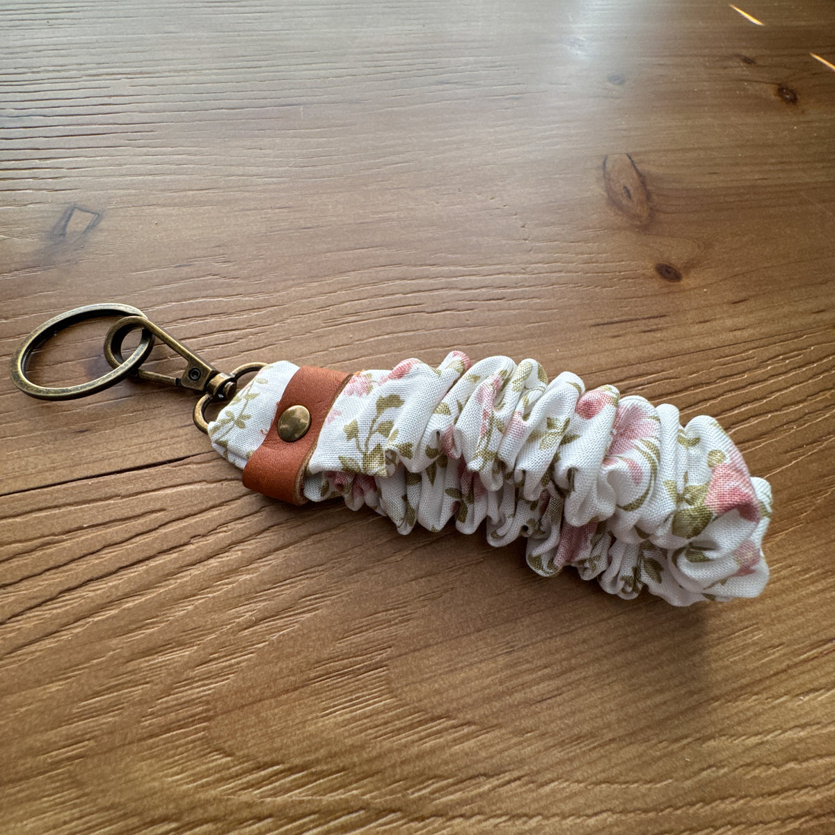 Handmade scrunchie wristlet keychain in cream floral fabric with leather accent and antique brass-tone hardware.