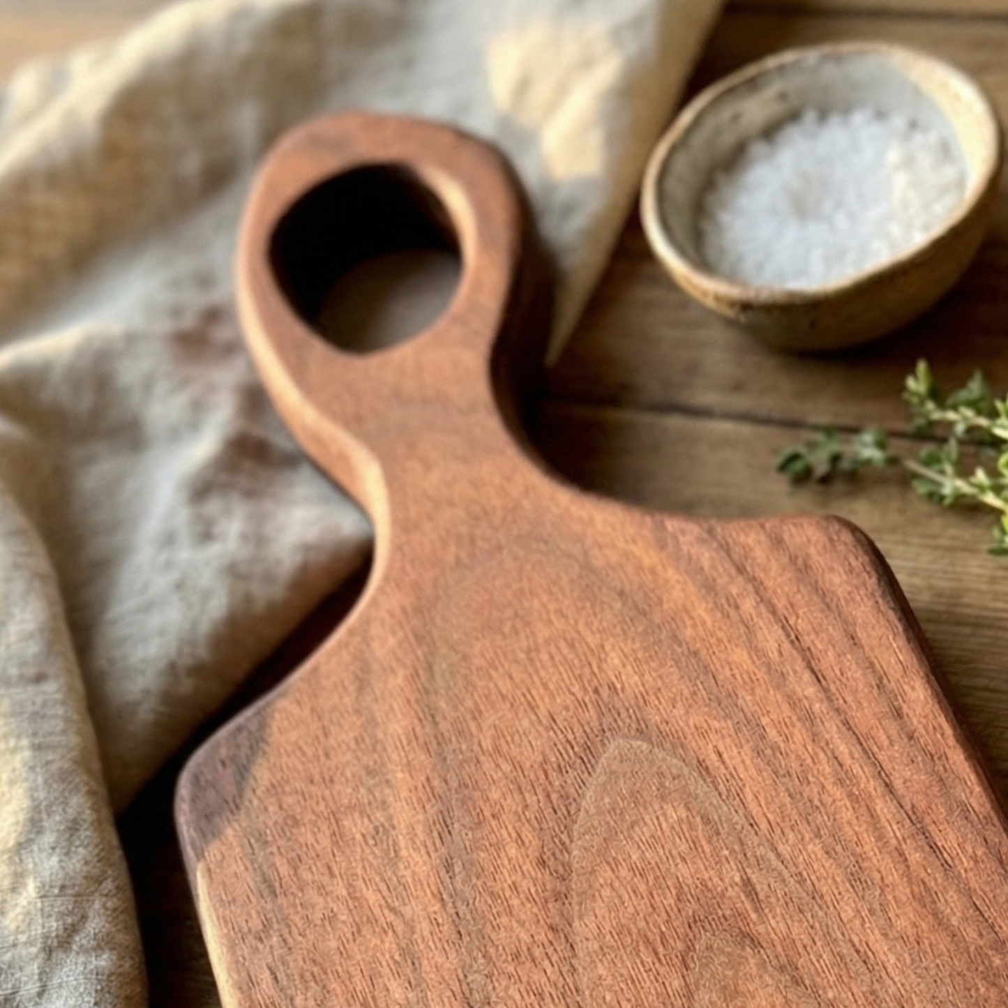 Handmade walnut snack size charcuterie board with handle on a rustic wood table styled with linen and sea salt