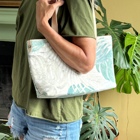 A person wearing an olive green shirt and blue jeans carries a large white tote bag with a light blue tropical leaf pattern and a beige shoulder strap, set against a background of green houseplants and a yellow wall.