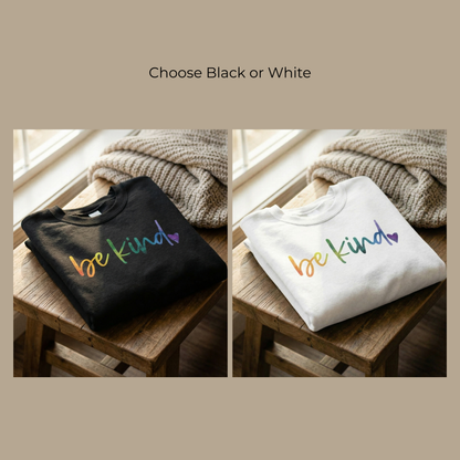 Two "Be Kind" rainbow crewneck sweatshirts in Midnight Black and Heirloom White are shown folded on a rustic wooden stool. The hand-lettered rainbow gradient and purple heart graphic are displayed clearly on both colors, highlighting the vibrant DTG print quality of this 1 Corinthians 16:14 inspired apparel.