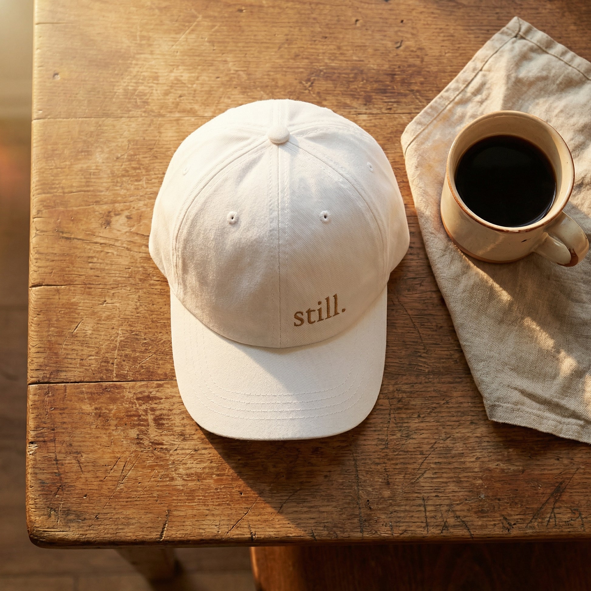 Minimalist white baseball cap embroidered with the word “still.” photographed with a coffee cup on a rustic wood table.