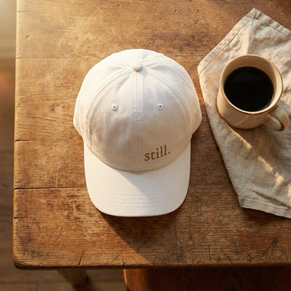 Minimalist white baseball cap embroidered with the word “still.” photographed with a coffee cup on a rustic wood table.