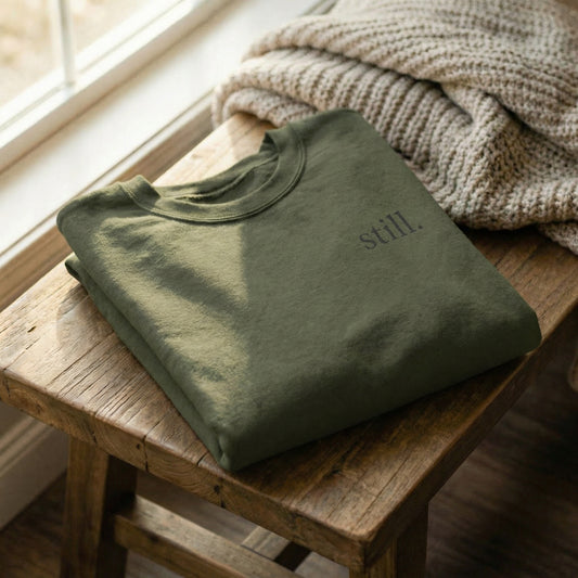 Folded green minimalist sweatshirt with “still.” typography on a wooden table with cozy knit texture.