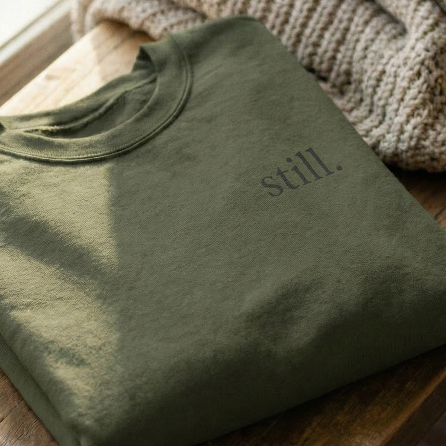 Folded green minimalist sweatshirt with “still.” typography on a wooden table with cozy knit texture.