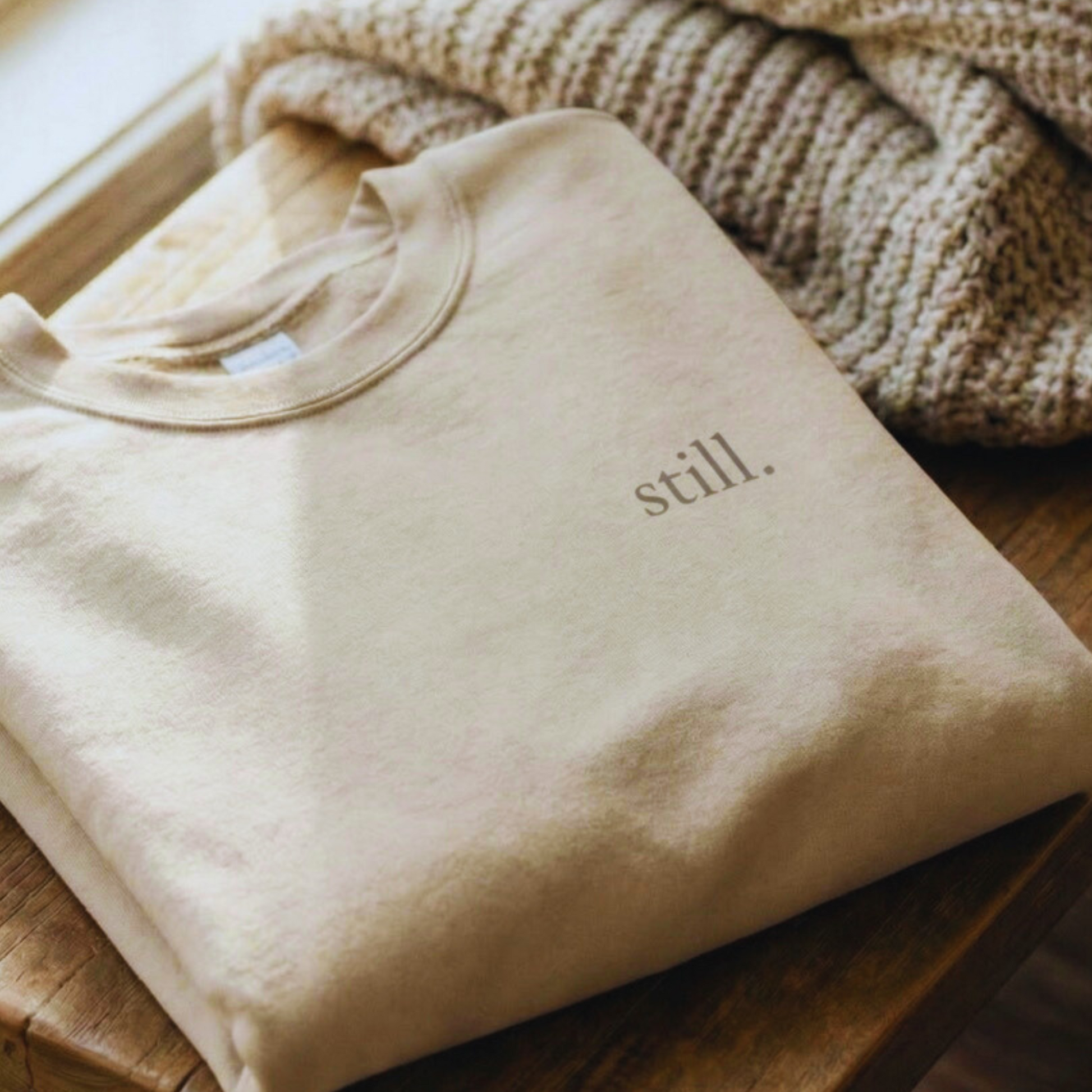 Folded sand-colored minimalist crewneck sweatshirt with the word “still.” on the chest, styled on a wooden stool beside a knit blanket.
