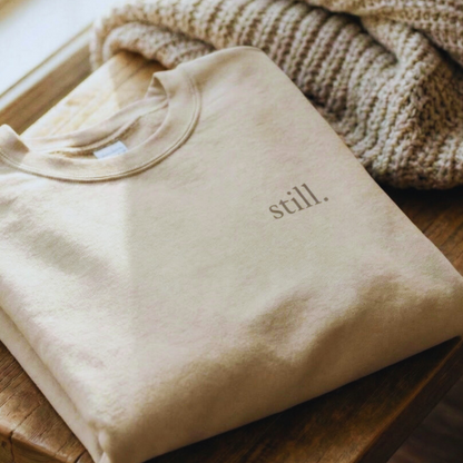 Folded sand-colored minimalist crewneck sweatshirt with the word “still.” on the chest, styled on a wooden stool beside a knit blanket.
