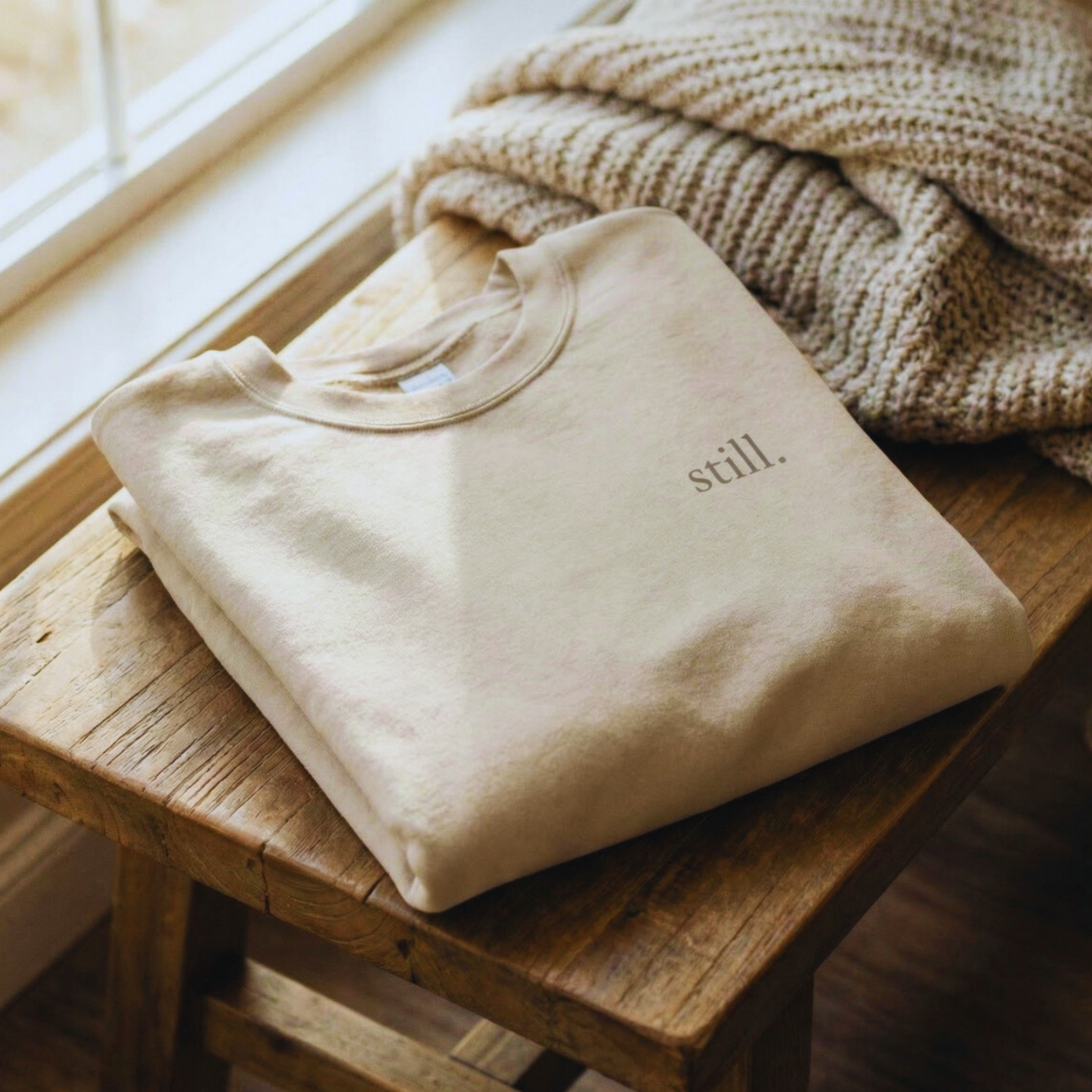 Folded sand-colored minimalist crewneck sweatshirt with the word “still.” on the chest, styled on a wooden stool beside a knit blanket.
