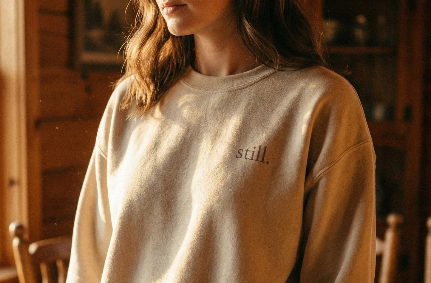 Minimalist neutral crewneck sweatshirt with the word “still.” printed on the chest in warm natural light.