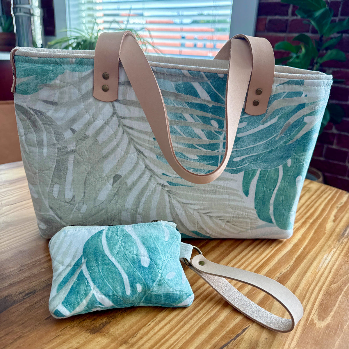 A large tote bag and matching small wristlet pouch with a tropical leaf print in teal, mint, and white, featuring tan faux-leather handles and wrist strap, displayed on a wooden table.