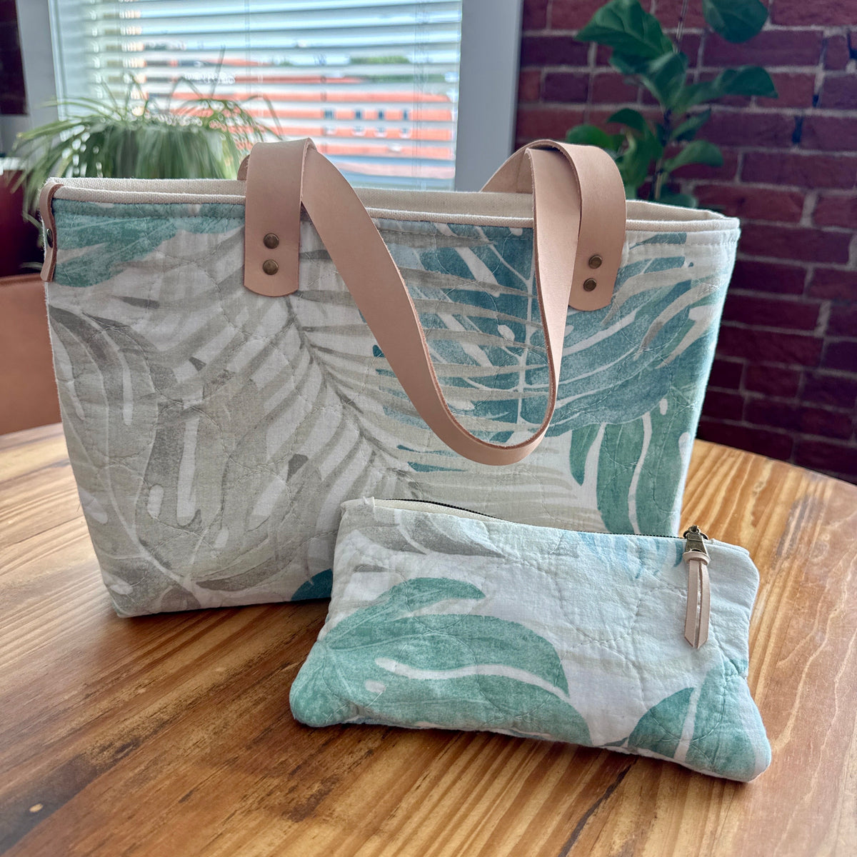 A matching fabric tote bag and two small wristlet pouches with a tropical leaf print in green and white, featuring light tan leather-look straps, displayed on a wooden table in a home setting.