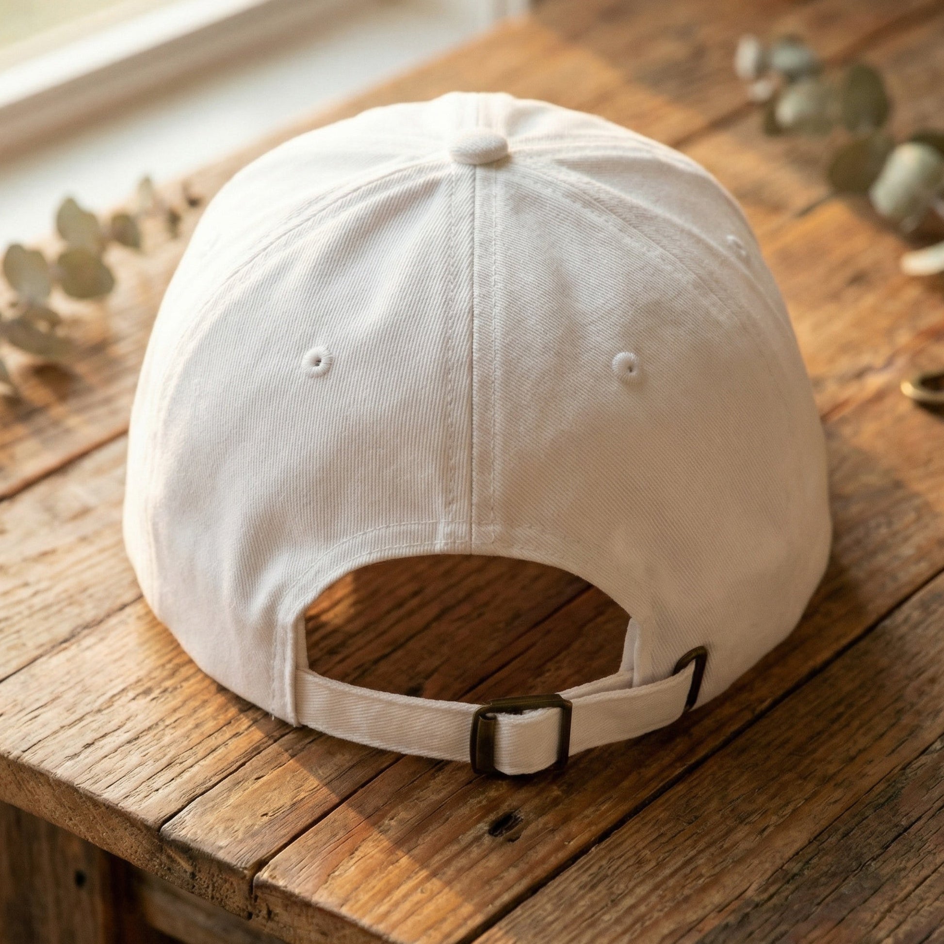 White embroidered baseball hat back view showing adjustable strap and antique brass buckle on wooden table