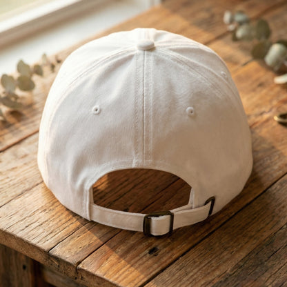 White embroidered baseball hat back view showing adjustable strap and antique brass buckle on wooden table