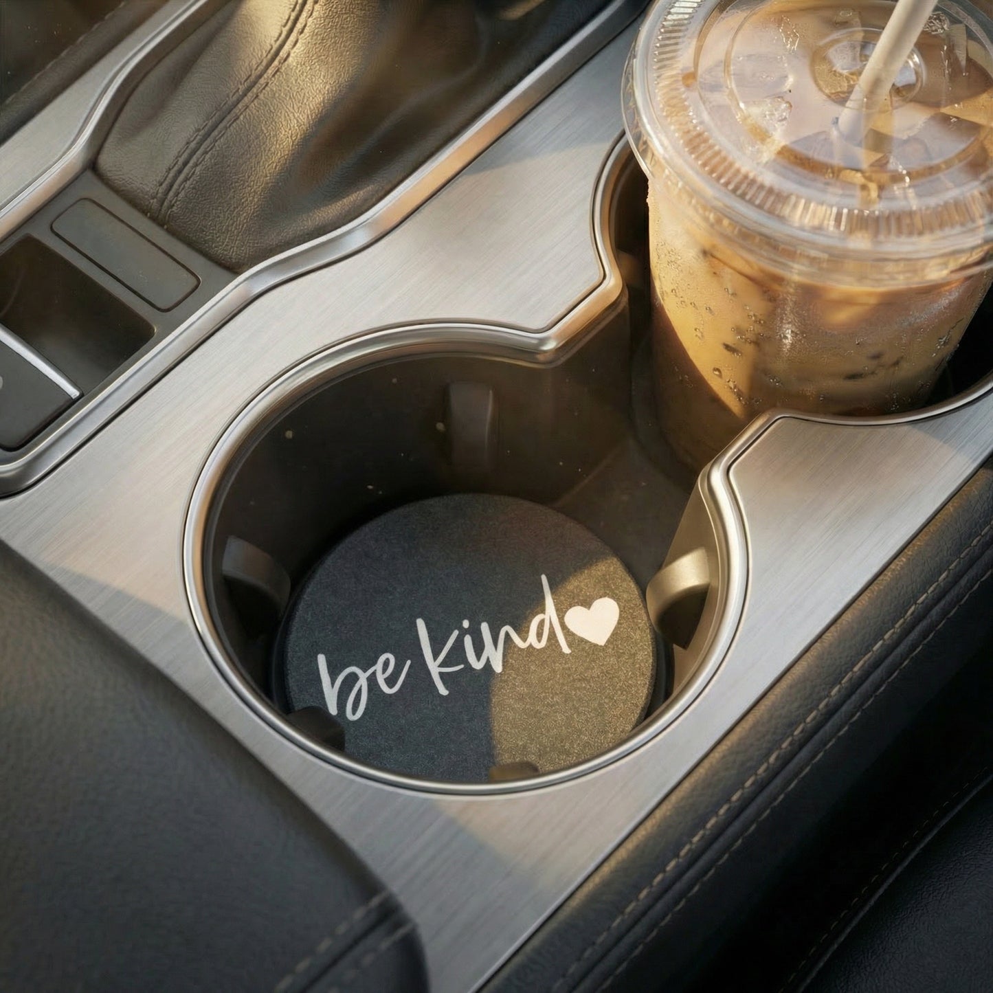 Car cup holder with a coaster labeled 'be kind' and an iced coffee drink.