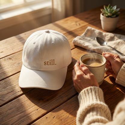 minimalist white “still” embroidered baseball hat on wooden table with iced coffee journal and glasses in warm morning light